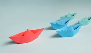 There are five origami boats placed on a blue background and lined in a row. First in the row is a red origami boat which is leading the others. In the second row there are two blue origami boats. Behind them are two green origami boats.